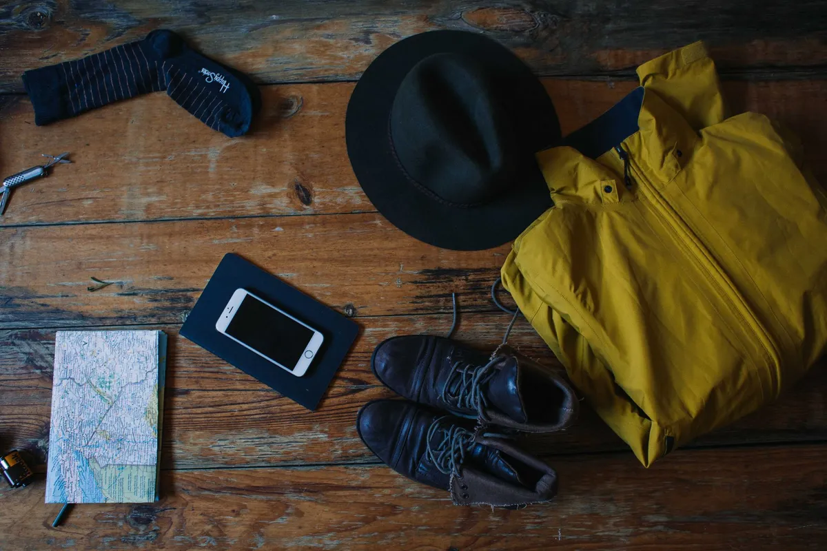 Travel essentials laid out on a wooden table, including a phone, a hat and a folded paper map